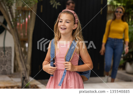 Smiling girl with backpack walking outside, parents following in background 135540885