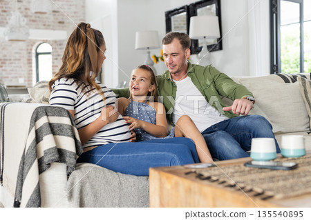 Family relaxing on sofa, young girl touching pregnant mother's belly, smiling warmly Family relaxing on sofa, young girl touching pregnant mother's belly, smiling warmly 135540895