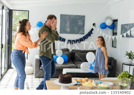 Family celebrating birthday surprise at home with balloons and cake, feeling joyful 135540902