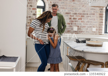Expectant mother and daughter embracing in kitchen while father watches lovingly 135540922