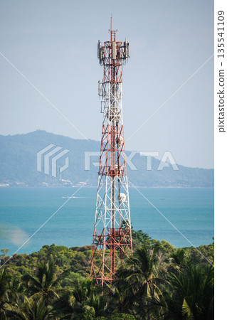 Tall mobile communication tower above tropical forest with ocean and island mountains in background under clear blue sky 135541109