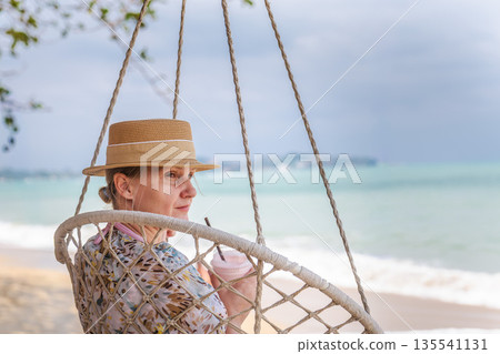 Middle aged woman relaxing in hanging chair on tropical beach, holding cold drink, ocean waves and cloudy sky in background, calm travel lifestyle Middle aged woman relaxing in hanging chair on tropical beach, holding cold drink, ocean waves and cloudy sky in background, calm travel lifestyle 135541131