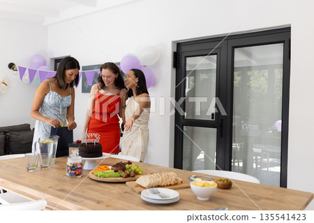 Laughing and chatting, three young women gather around chocolate cake at home 135541423