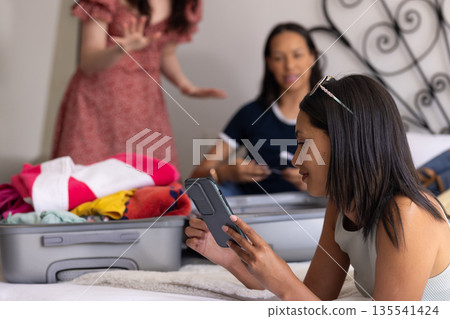 Packing suitcase on bed, young women with diverse backgrounds chat happily Packing suitcase on bed, young women with diverse backgrounds chat happily 135541424