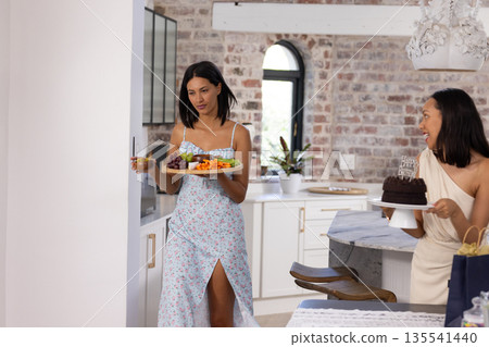 Carrying platter of colorful fruits and vegetables, two Asian women in stylish dresses smile warmly 135541440