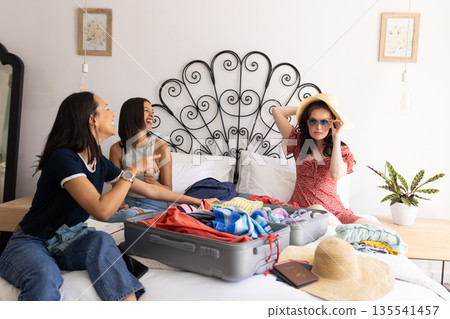 Packing suitcase on bed, three young women are preparing for trip Packing suitcase on bed, three young women are preparing for trip 135541457