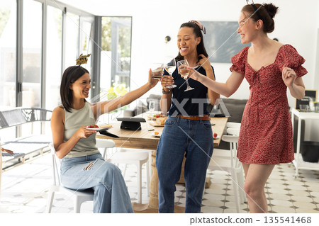 Laughing and toasting with wine glasses, three young women enjoy sunny afternoon indoors 135541468