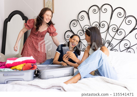 Packing suitcase on bed, three young women are laughing together Packing suitcase on bed, three young women are laughing together 135541471