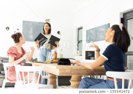 Holding menus and smiling, young woman with Birthday Girl headband stands by table Holding menus and smiling, young woman with Birthday Girl headband stands by table 135541472
