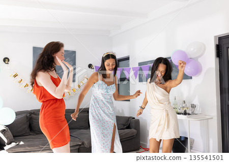 Dancing joyfully in bright living room, three young women celebrate together 135541501