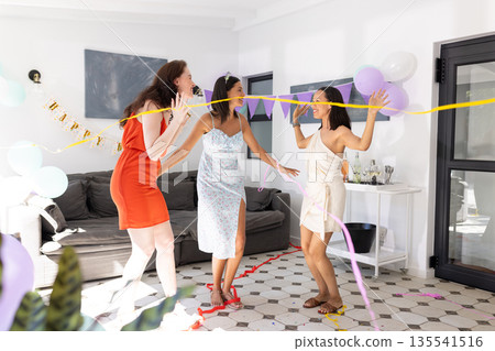 Dancing joyfully, three women in colorful dresses celebrate indoors with balloons and streamers Dancing joyfully, three women in colorful dresses celebrate indoors with balloons and streamers 135541516