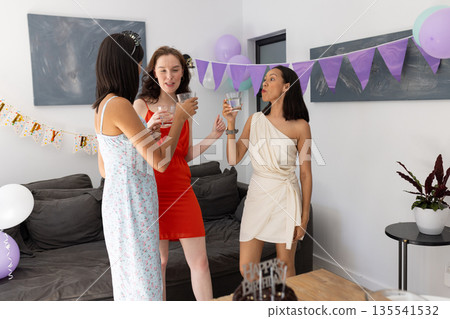 Three young women celebrating at home, wearing colorful dresses and holding drinks 135541532