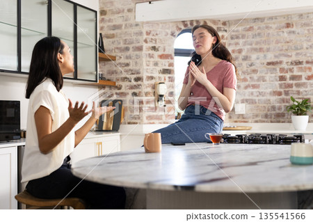 Engaging in lively conversation, two young women sit in modern kitchen 135541566