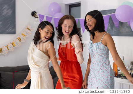 Dancing joyfully, three young women in colorful dresses celebrate at lively party 135541580