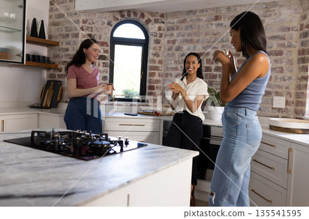 Laughing and chatting, three young women gather in cozy kitchen Laughing and chatting, three young women gather in cozy kitchen 135541595