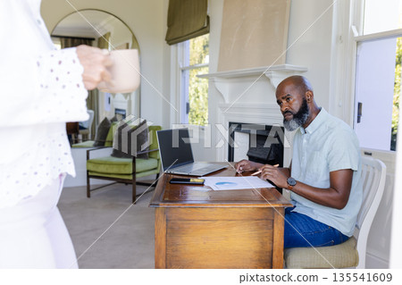 Man working at home office desk with laptop, receiving coffee from woman 135541609