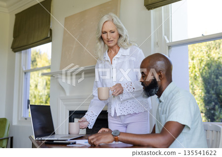 Mature woman serving coffee to man working on laptop at home office 135541622