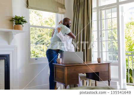Couple embracing warmly at home, laptop and coffee mugs on wooden desk Couple embracing warmly at home, laptop and coffee mugs on wooden desk 135541649