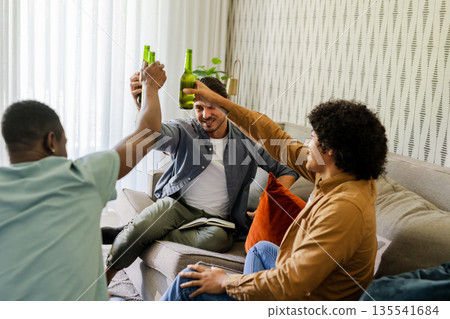 Friends celebrating with beer bottles on couch, enjoying joyful moment together 135541684