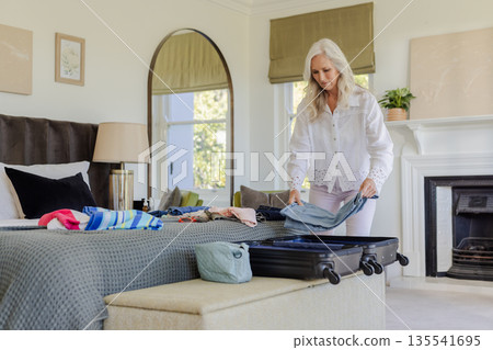 Packing suitcase, mature woman preparing for trip in cozy bedroom Packing suitcase, mature woman preparing for trip in cozy bedroom 135541695