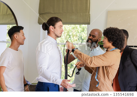 Groom getting ready with friends helping tie necktie in bright room Groom getting ready with friends helping tie necktie in bright room 135541698