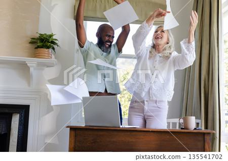 Joyful couple celebrating success, throwing papers in air near laptop at home Joyful couple celebrating success, throwing papers in air near laptop at home 135541702