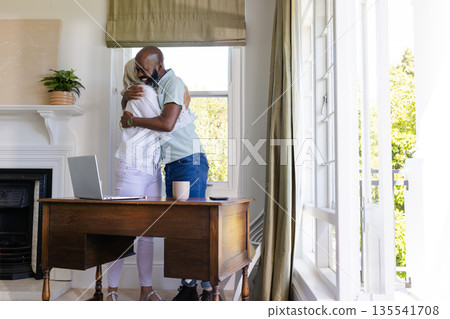 Couple embracing warmly by window, laptop and coffee cup on wooden desk 135541708