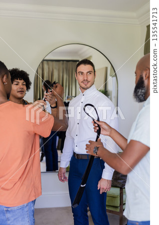 Groom preparing for wedding day with friends helping adjust tie in bedroom Groom preparing for wedding day with friends helping adjust tie in bedroom 135541713