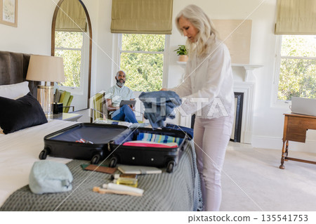 Woman packing suitcase in bedroom while man sitting on bed, preparing for trip Woman packing suitcase in bedroom while man sitting on bed, preparing for trip 135541753