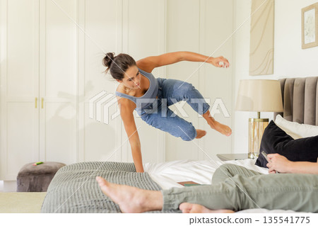 Woman playfully jumping on bed in casual jeans and tank top at home Woman playfully jumping on bed in casual jeans and tank top at home 135541775