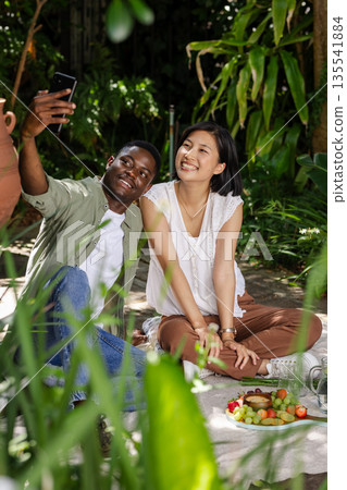 Couple enjoying picnic, taking selfie together, surrounded by lush greenery outdoors Couple enjoying picnic, taking selfie together, surrounded by lush greenery outdoors 135541884