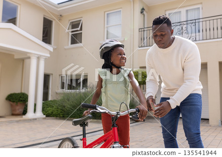 Father teaching daughter to ride bicycle in driveway, both smiling and engaged 135541944