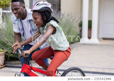 Father teaching daughter to ride bicycle in driveway, both smiling and excited 135541946