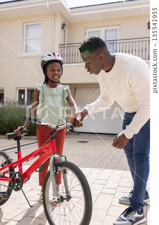 Father teaching daughter to ride bicycle in driveway, both smiling happily Father teaching daughter to ride bicycle in driveway, both smiling happily 135541955