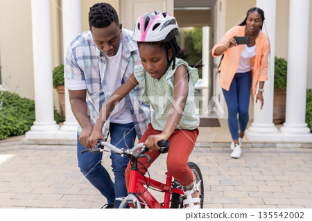 Teaching daughter to ride bicycle, African American father while mother captures moment 135542002