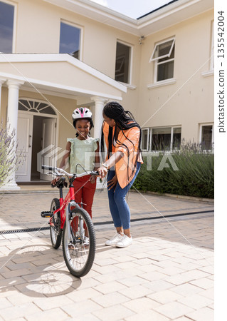 Teaching daughter to ride bicycle, African American mother outside family home smiling 135542004