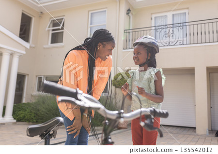 Mother and daughter smiling, preparing for bike ride in front of house 135542012