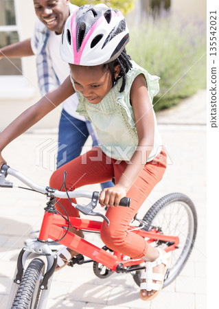 Father supporting African American child riding bicycle outdoors, both smiling Father supporting African American child riding bicycle outdoors, both smiling 135542021