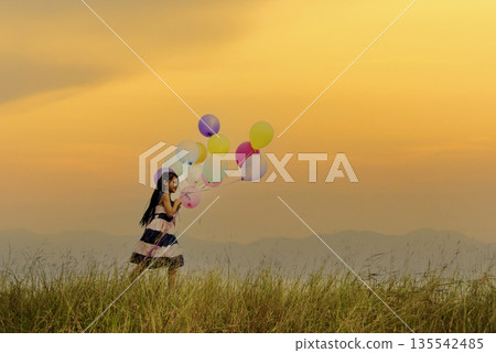 Cheerful cute girl holding balloons running on green meadow sunrise golden vibrant sky happiness. Hands holding vibrant air balloons play on birthday party happy times summer on gold sky outdoor Cheerful cute girl holding balloons running on green meadow sunrise golden vibrant sky happiness. Hands holding vibrant air balloons play on birthday party happy times summer on gold sky outdoor 135542485
