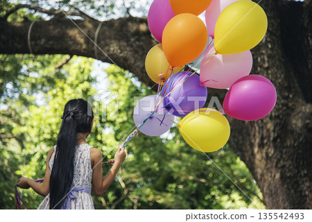 Cheerful cute girl holding balloons running on green meadow white cloud and blue sky with happiness. Hands holding vibrant air balloons play on birthday party happy times summer on sunlight outdoor 135542493