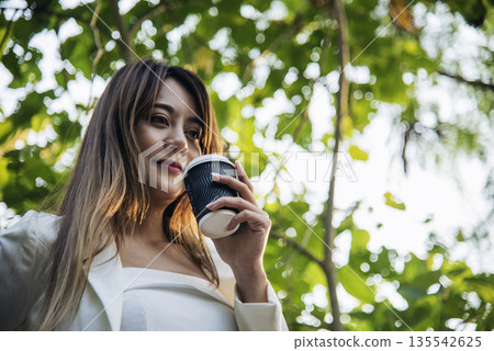 Woman drinking coffee hands holding hot disposable cup in green park. Happy Relax beautiful asian woman smiling face standing outdoors garden. Young women enjoy nature morning Freedom Lifestyle. 135542625