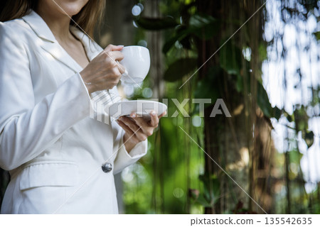 Woman drinking coffee hands holding hot disposable cup in green park. Happy Relax beautiful asian woman smiling face standing outdoors garden. Young women enjoy nature morning Freedom Lifestyle. 135542635