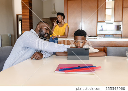 Father helping son with tablet at home while mother talks on phone nearby Father helping son with tablet at home while mother talks on phone nearby 135542968