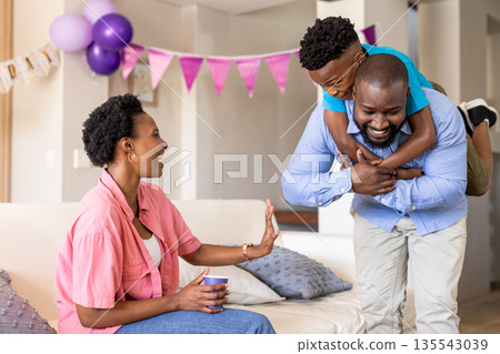 Father playfully carrying son on back while mother laughs on cozy living room sofa 135543039