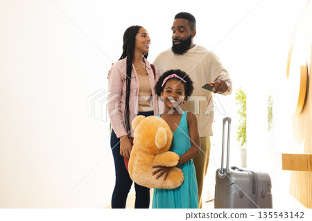 Family arriving at hotel, young girl holding teddy bear, parents smiling warmly 135543142