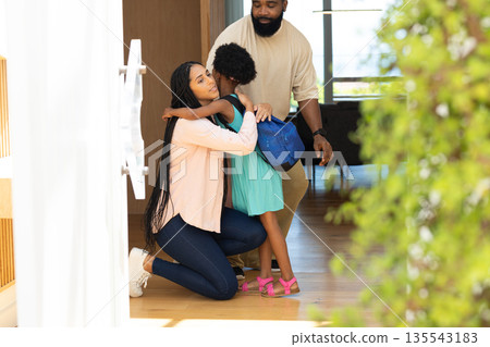 Mother hugging daughter with backpack while father smiling at home entrance 135543183
