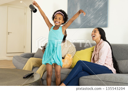 Joyful girl in blue dress dancing at home with family smiling on couch 135543204