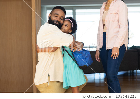 Father hugging daughter with backpack, mother smiling nearby in cozy living room Father hugging daughter with backpack, mother smiling nearby in cozy living room 135543223