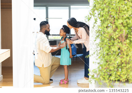 Parents helping daughter with backpack, preparing for school at home, smiling warmly Parents helping daughter with backpack, preparing for school at home, smiling warmly 135543225