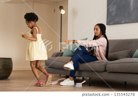 Mother sitting on sofa pointing while daughter playfully running in living room Mother sitting on sofa pointing while daughter playfully running in living room 135543278